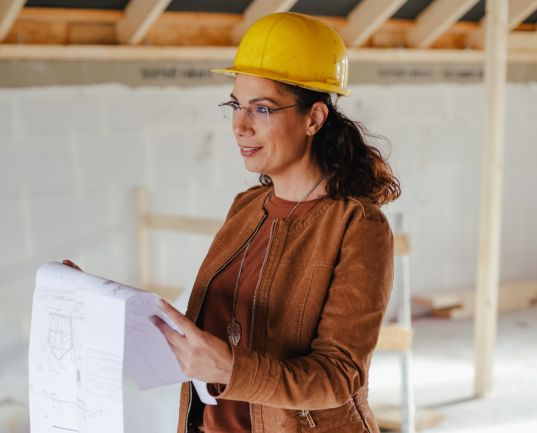 Eine Frau in gelber Bauhelme und Brille hält Baupläne in einer Baustelle.